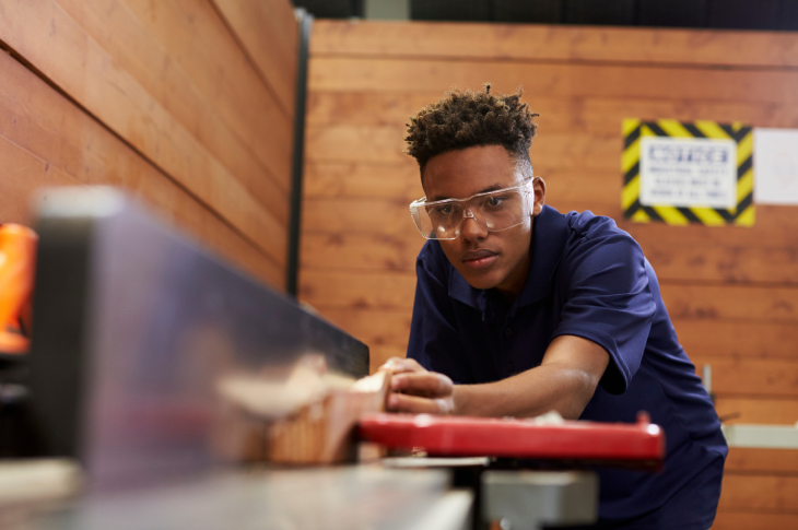A student in the SAIT carpentry program wearing personal protective equipment saws a piece of wood on a table.