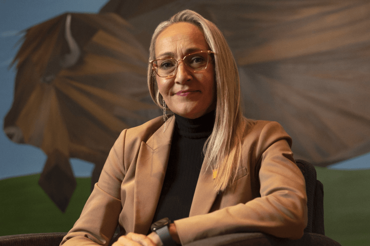 A headshot of Jennifer Russell, Director, Indigenous Engagement. She wears a beige suite jacket over a black turtleneck and sits in front of a mural of a buffalo in Natoysopoyiis.