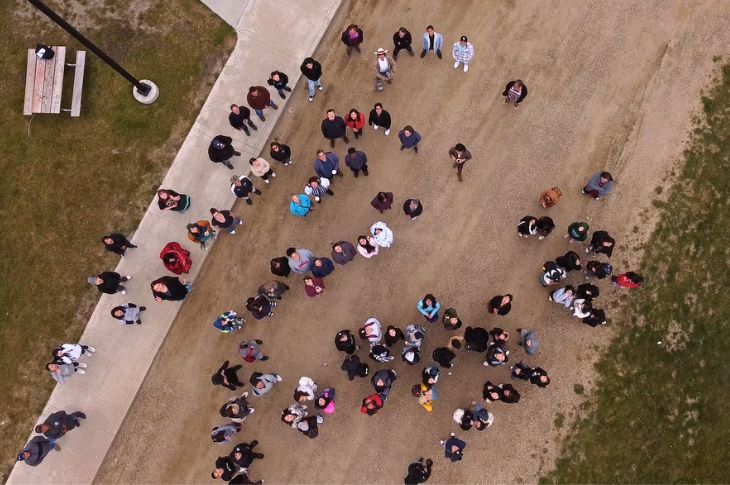 About 50 people stand on a road and look up at the camera.