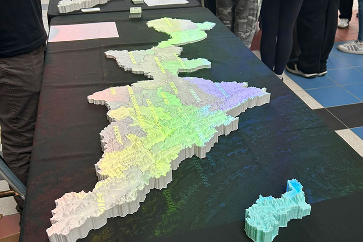 A large white 3D map model showing topography in ridges and valleys is overlaid with a colour computer projection map.  