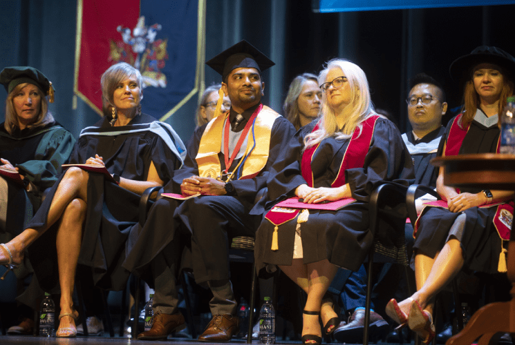 Sanoop Sadique wearing his cap and gown sits on stage during a graduation ceremony, surrounded by members of SAIT"s faculty.