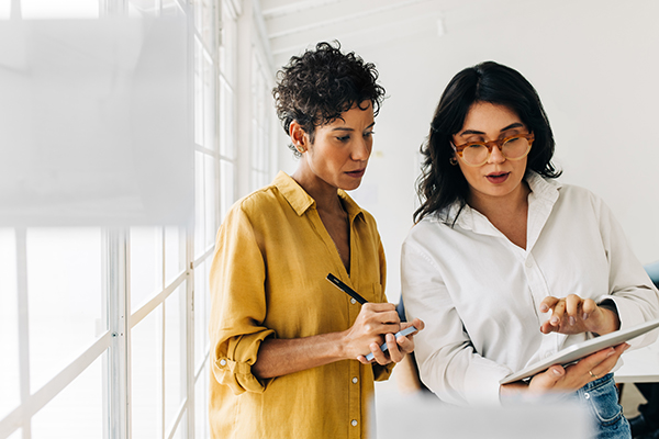 Two women discussing while one is writing notes down and the other one is pointing to her iPad.