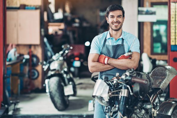 A motorcycle mechanic apprentice crosses his arms and smiles at the camera.
