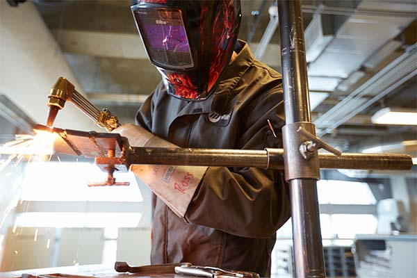 Sparks fly as a welder apprentice users a welding torch on piping. They are wearing a welding helmet, safety gloves and coveralls.